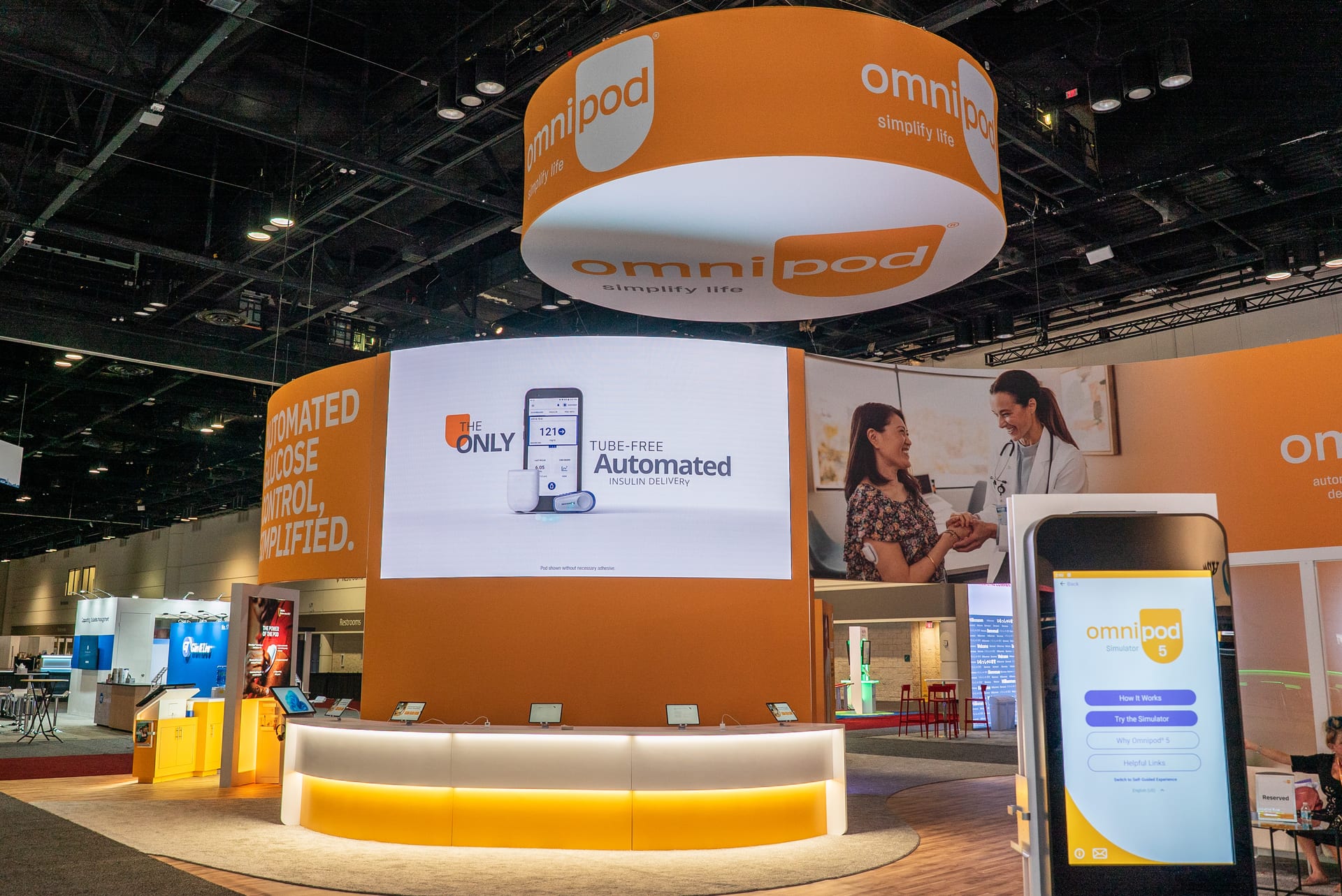 The Omnipod booth at ADA 2024, its LED wall running tube-free insulin delivery content over a lit reception counter, with the simulator kiosk standing at the aisle