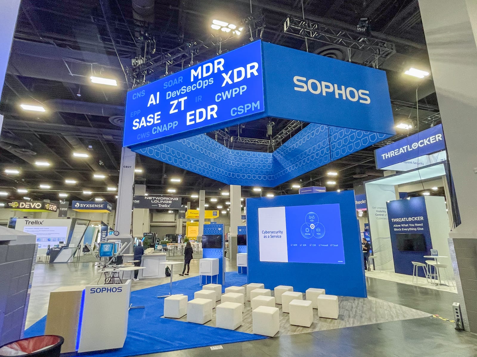 A glowing blue Sophos overhead LED sign, one face listing security acronyms, above a Cybersecurity as a Service LED wall and white seating on the show floor