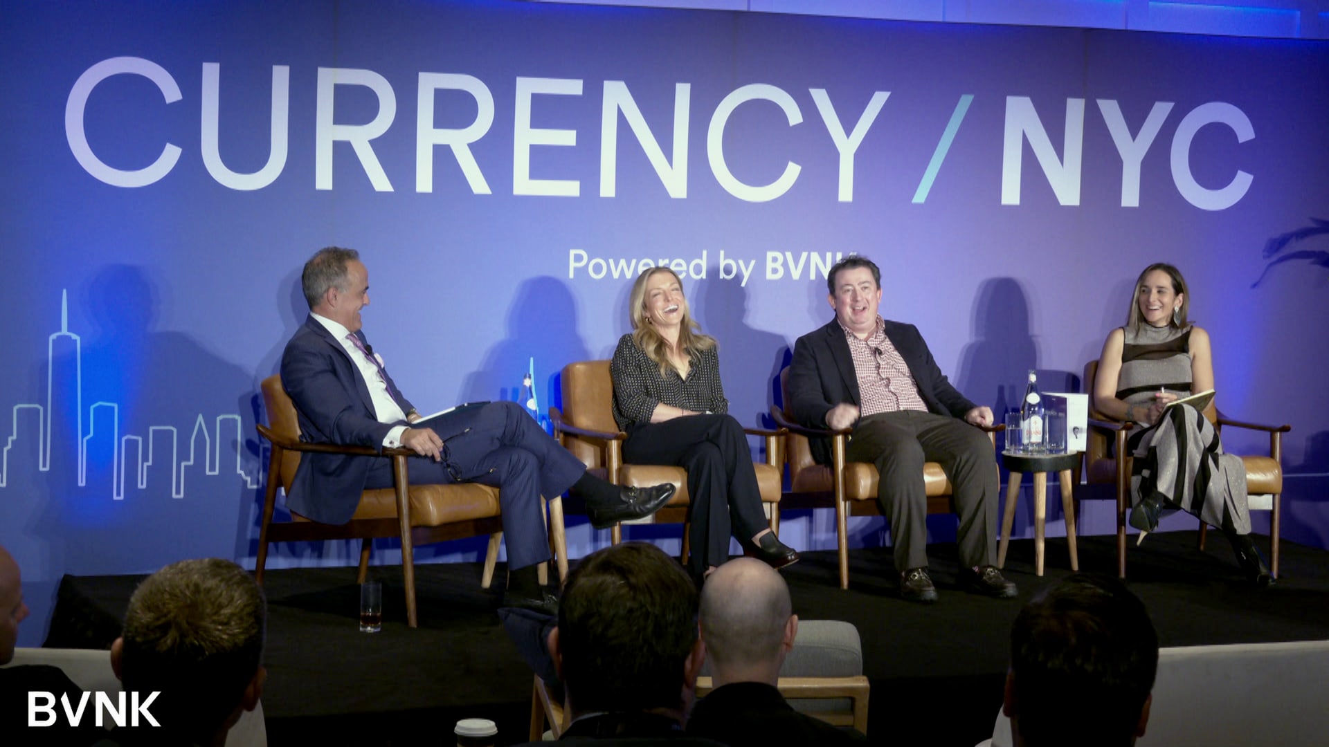 A four-person panel on stage at BVNK's Currency NYC, lit against a blue Currency / NYC backdrop with a city skyline, the audience in silhouette up front