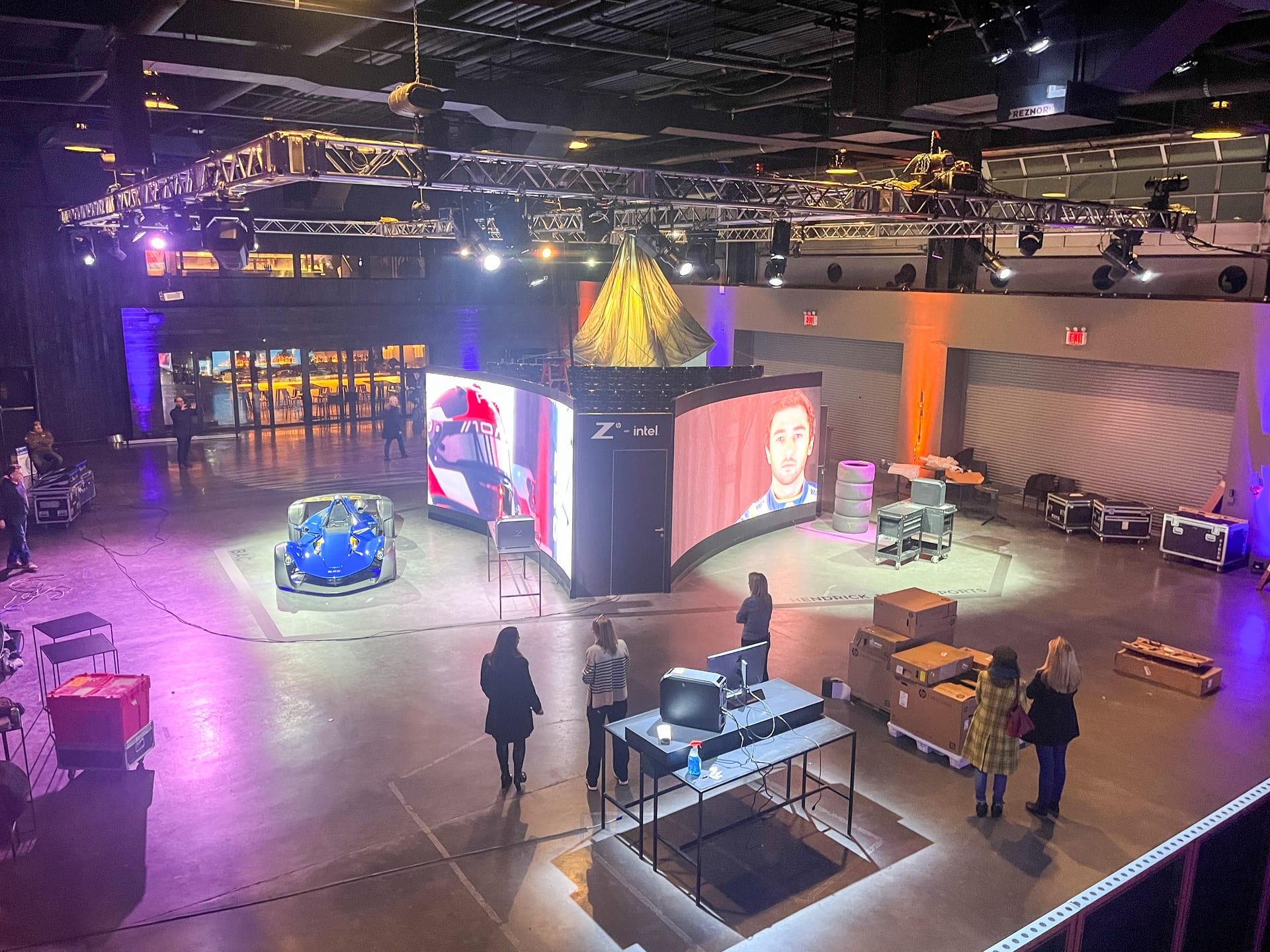 The Z by HP launch floor from above: three curved LED walls, the blue BAC car, workstation carts, and guests moving between the worlds under the pier's truss