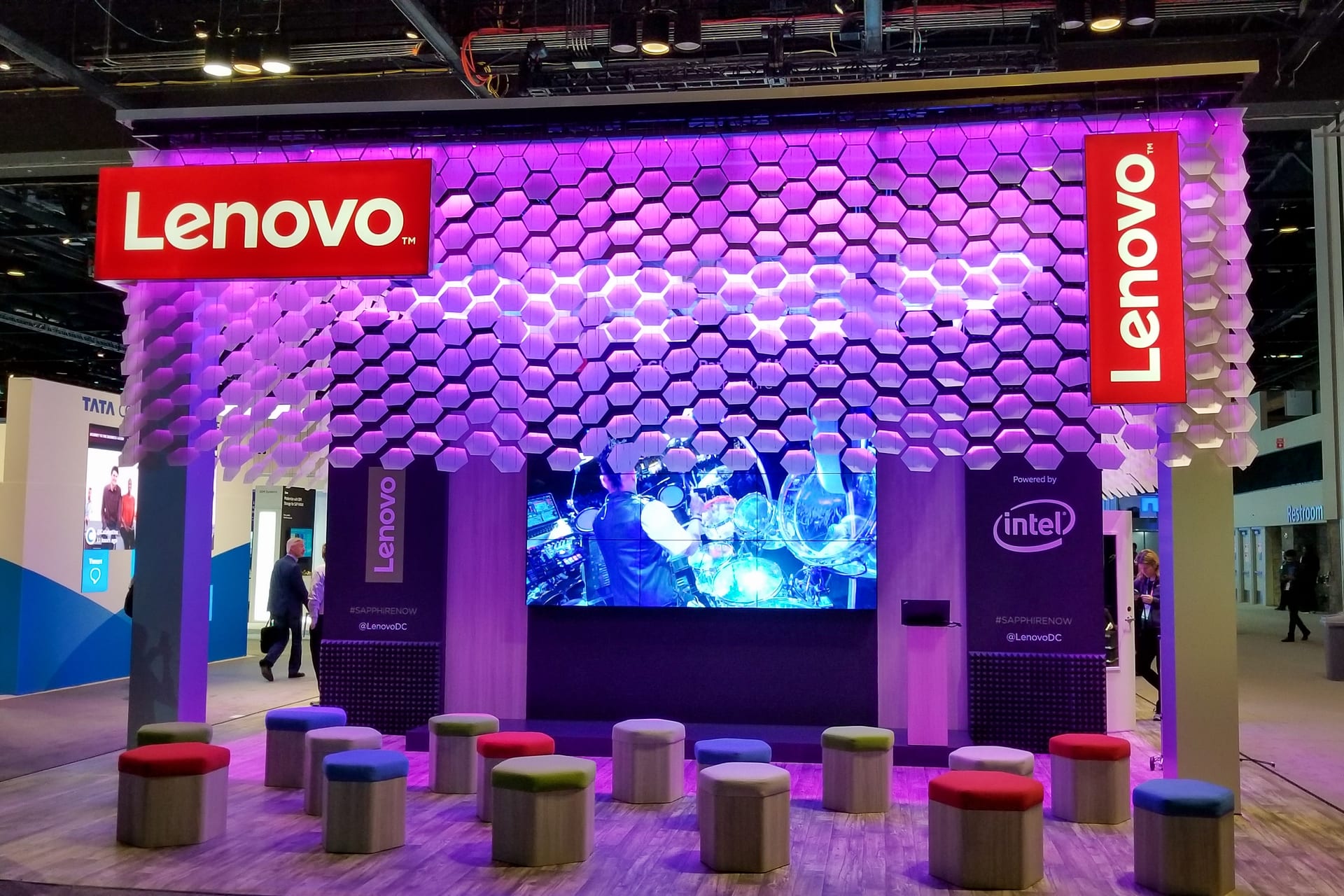 The Lenovo theater at SAPPHIRE 2019 before doors: a canopy of hexagonal cells lit magenta over the nine-panel video wall, with hexagonal ottomans set out for the audience
