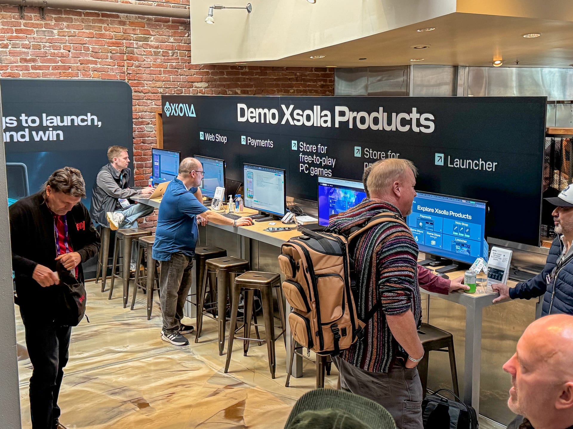 Developers working through Xsolla's products at the Clubhouse demo bar, a row of monitor stations and stools along an exposed-brick wall