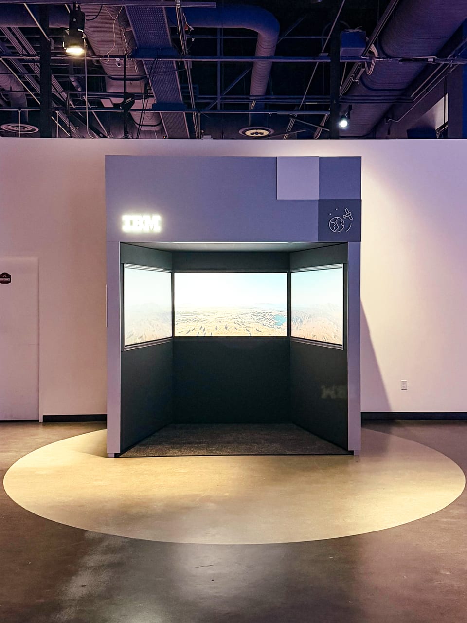 A walk-in alcove exhibit with a lit IBM logo above the entrance, three touchscreens inside running one continuous landscape view