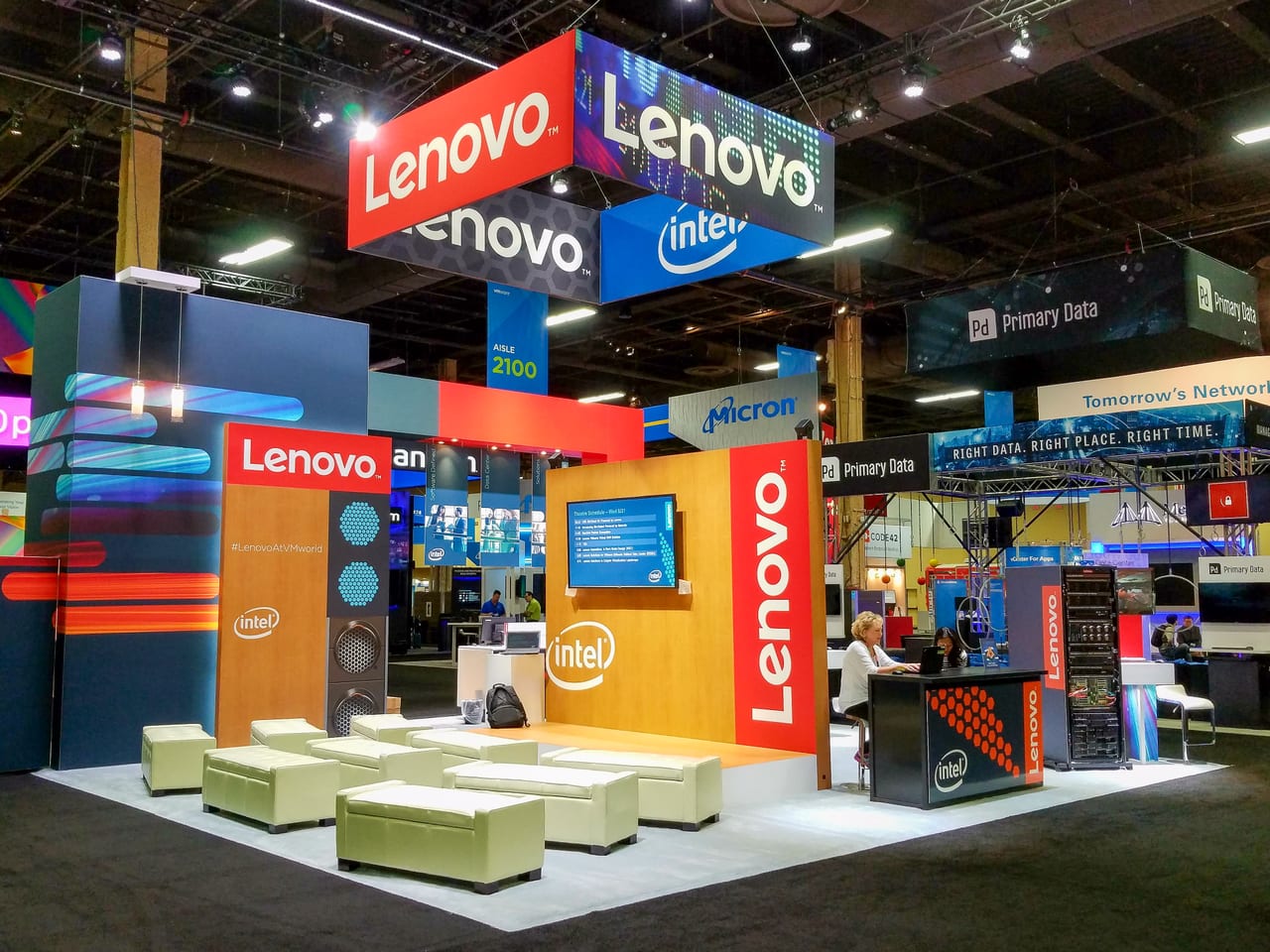The Lenovo and Intel booth at VMworld 2016 before doors: wood theater stage with the day's schedule on screen, cream ottomans, red Lenovo towers, and the hanging sign overhead