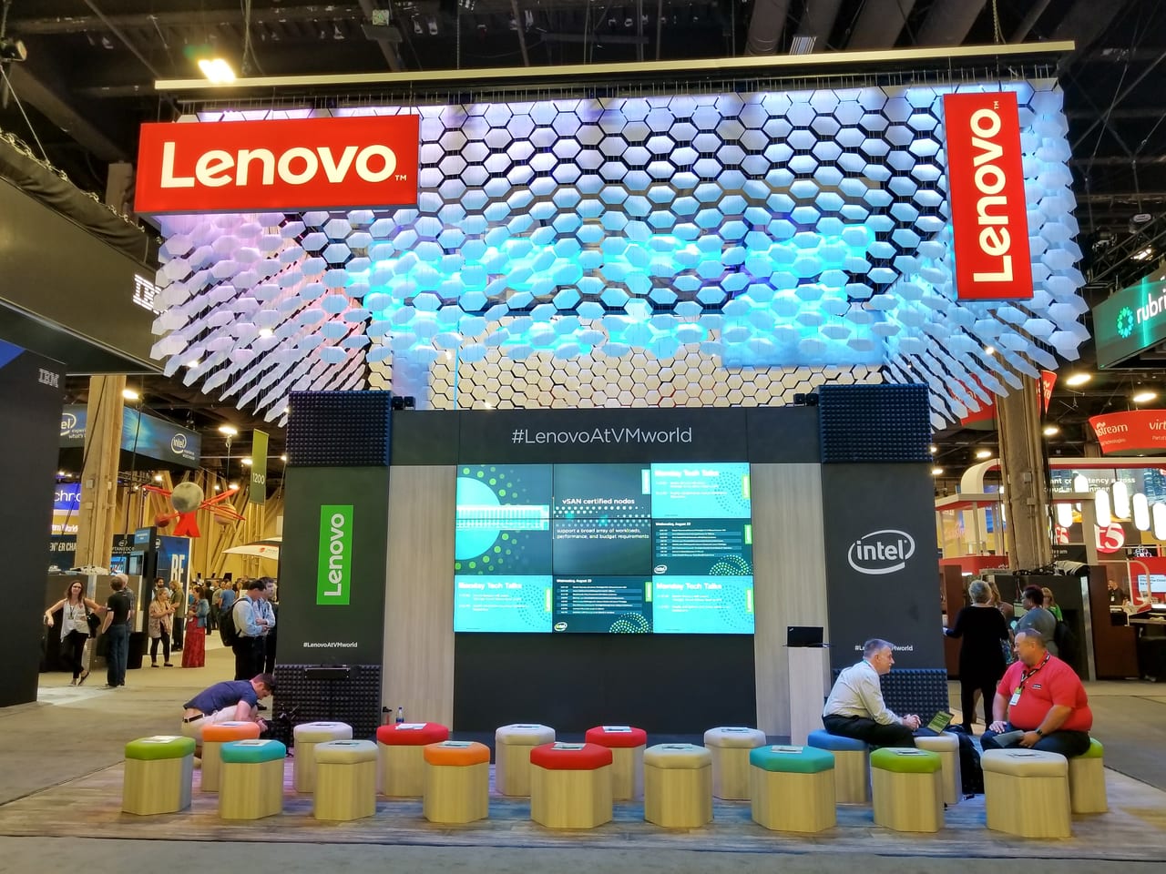 Lenovo's VMworld 2018 booth before the crowds: a canopy of hanging hexagonal cells lit blue and purple over a nine-panel video wall and hex-shaped stools