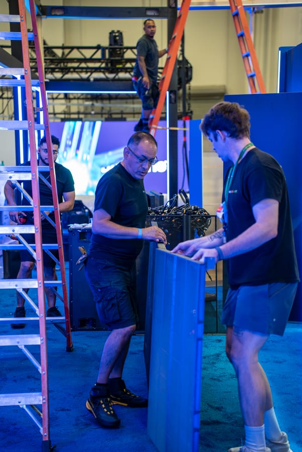 Two crew members handing off a piece of gear at a blue counter during load-in, ladders and rigging behind them