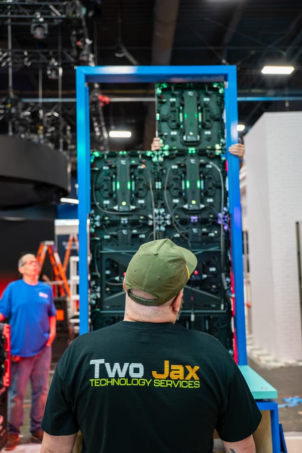 A crew member in a Two Jax shirt facing a stack of LED tiles in its support frame during assembly