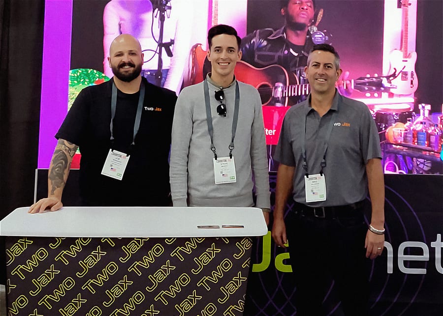 Three crew members at the Two Jax booth at Exhibitor Live, a branded counter in front of them and an LED wall running concert footage behind