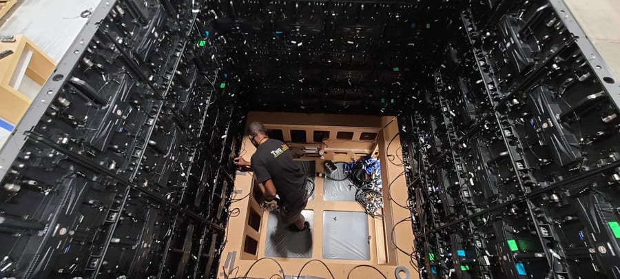 A technician in a Two Jax shirt working inside a room lined floor to ceiling with the backs of LED tiles, cabling run to a plywood counter