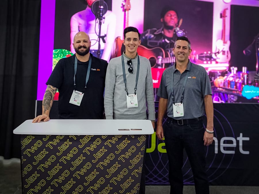 Three crew members at the Two Jax booth at Exhibitor Live, a branded counter in front of them and an LED wall running concert footage behind